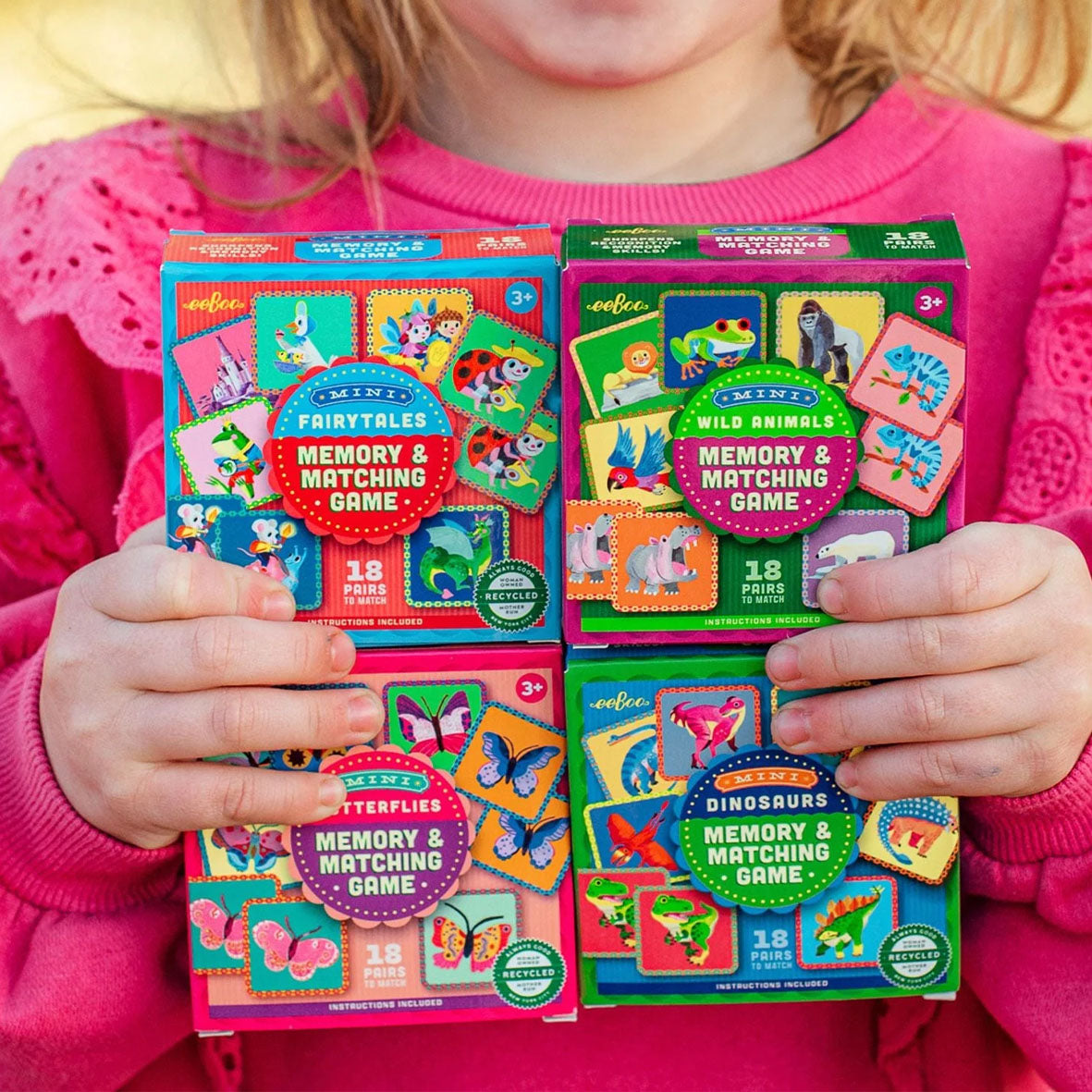 A child holding four mini boxed memory and matching games. The boxes are labeled 'Memory & Matching Game' by the brand eeBoo and show different themed sets: Fairytales, Wild Animals, Butterflies, and Dinosaurs.