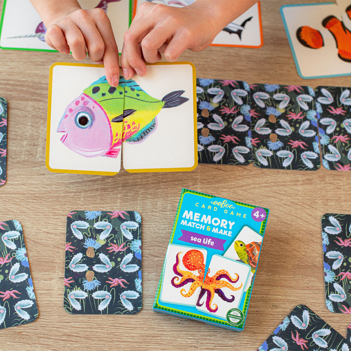 Child playing with a colourful sea life card game, matching a fish card to its patterned card and the box for 'Memory Match & Make sea life' card game by eeboo.