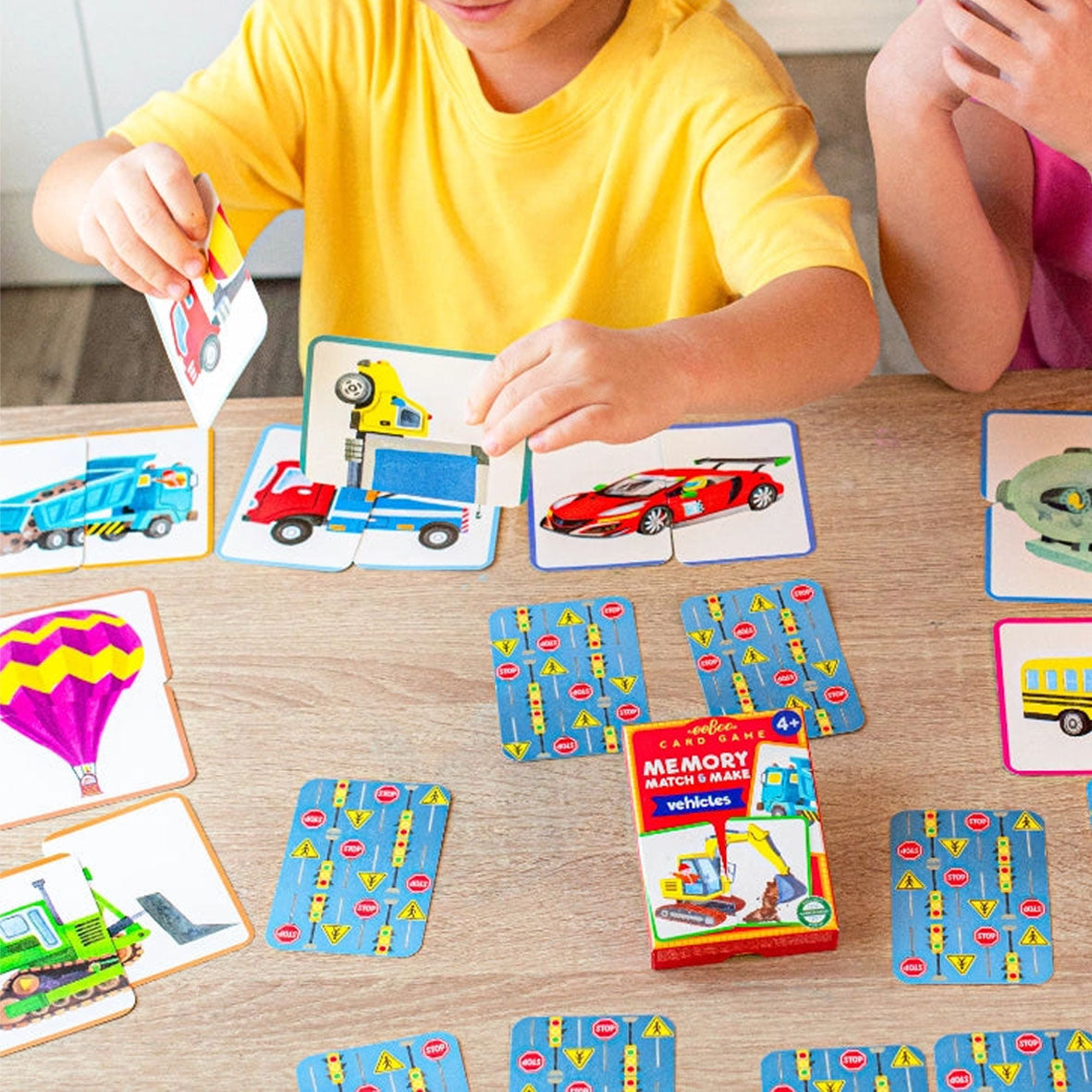 Children playing with a Memory Match Card with cards showing colourful vehicles, a hot air balloon, and construction trucks. The product box is visible in the center, and the brand 'Memory Match Card and Make Vehicles' is on the packaging.